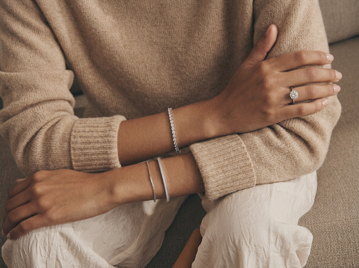 Crossed arms in cream silk wearing a sterling silver moissanite tennis bracelet, stacked silver bracelets, and a solitaire moissanite ring in warm natural light